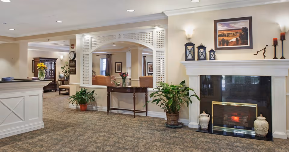 Interior view of a senior living facility lobby area with a white reception desk on the left, a decorative wooden table with flowers in the center, and a fireplace with candles and framed artwork on the right. The space is carpeted and decorated with potted plants and wall sconces.