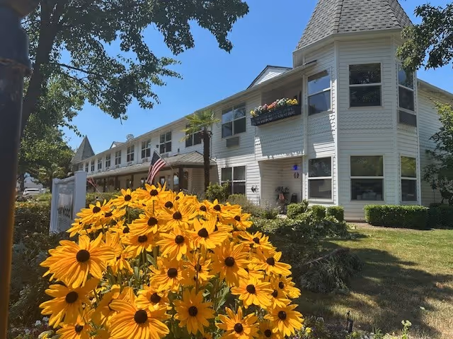 Exterior view of a two-story white building with multiple windows and flower boxes, surrounded by greenery and a large cluster of yellow flowers in the foreground under a clear blue sky.
