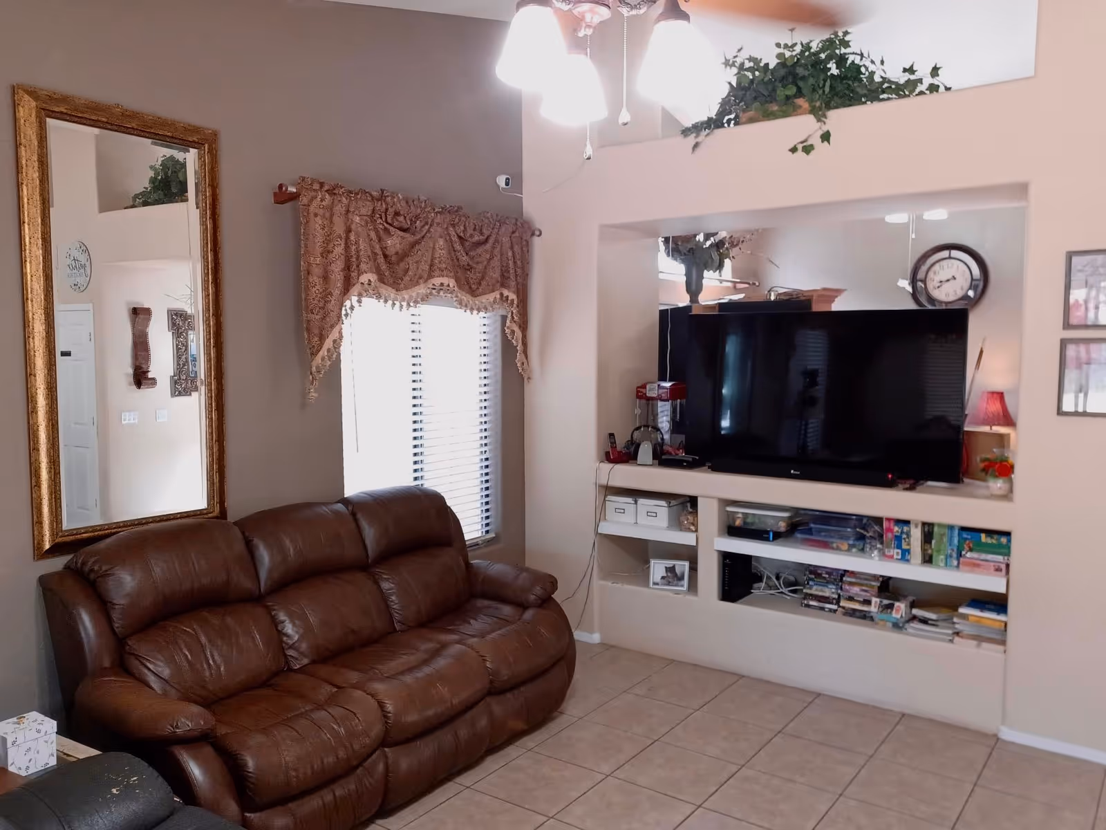 A cozy living room with a brown leather sofa, a large wall-mounted flat-screen TV, and built-in shelves below the TV holding various items including board games and books. There is a large mirror with a gold frame on the left wall, a window with decorative curtains, and a ceiling light fixture with multiple bulbs. A plant is placed on a ledge above the TV area.