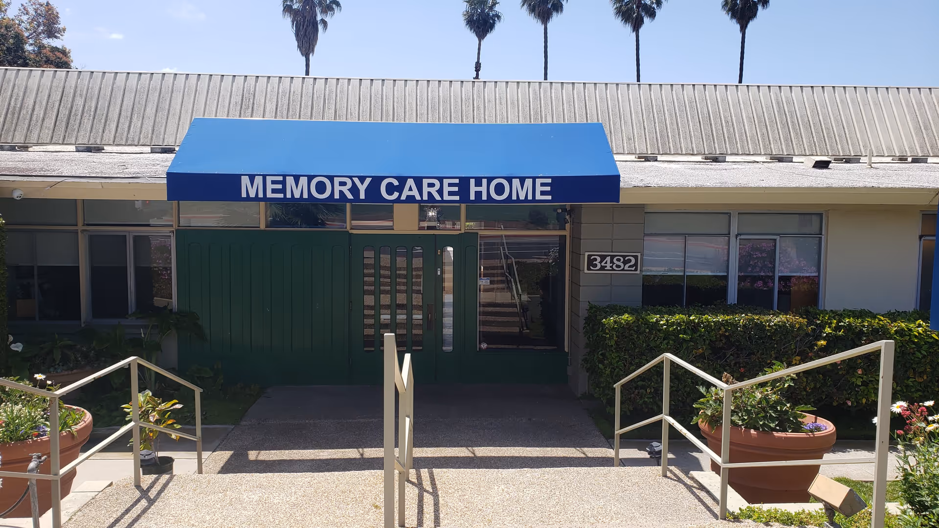 Entrance to a memory care home with a blue awning that reads 'MEMORY CARE HOME'. There are green double doors beneath the awning, a concrete pathway with handrails leading up to the entrance, large potted plants on either side, and the building number 3482 displayed on the wall to the right. Palm trees are visible in the background.