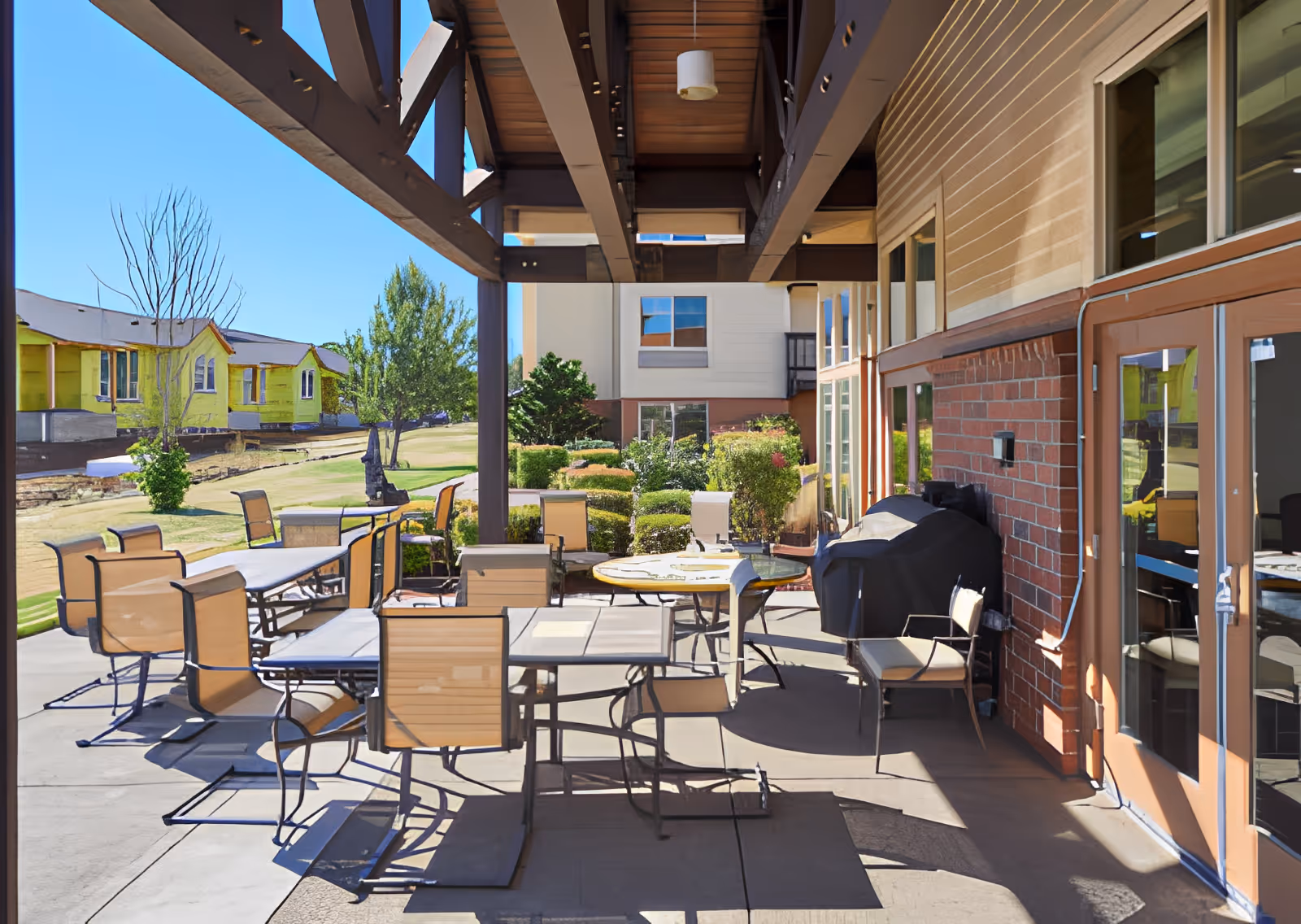Outdoor patio area at Elliott Residence with multiple tables and chairs arranged under a wooden pergola. There is a barbecue grill against the brick wall of the building, and the patio overlooks a grassy area with trees and other buildings in the background under a clear blue sky.