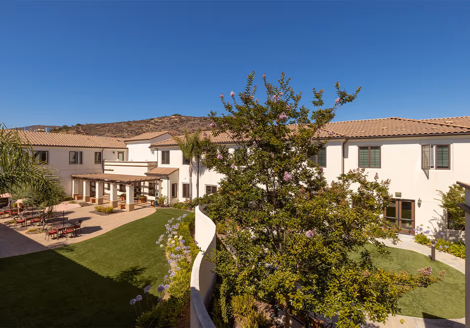 Outdoor courtyard area of Westmont at San Miguel Ranch featuring a well-maintained lawn, flowering trees and plants, a pergola with seating, and a two-story building with beige walls and tiled roof under a clear blue sky.