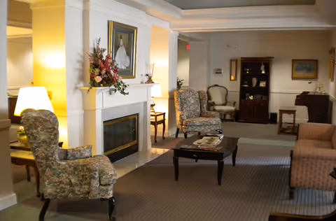 A cozy common room with floral upholstered armchairs, a fireplace, coffee table, and warm lamps.