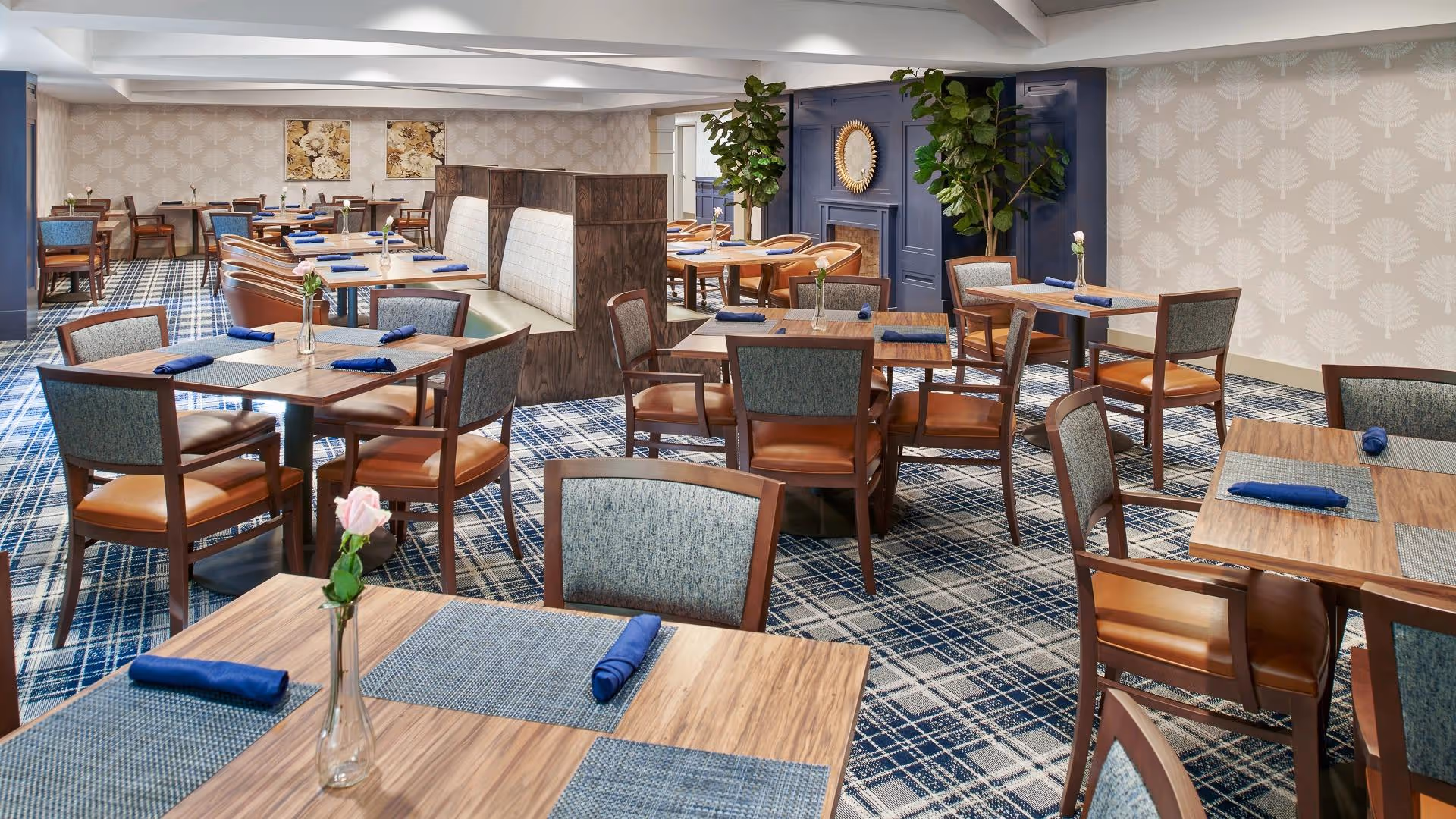 A well-lit dining room with multiple wooden tables and chairs arranged neatly. Each table has a blue placemat and a rolled blue napkin, with a small vase holding a single flower. The room features patterned carpet flooring, wallpaper with a subtle leaf design, and decorative plants near a blue accent wall with a mirror above a fireplace.