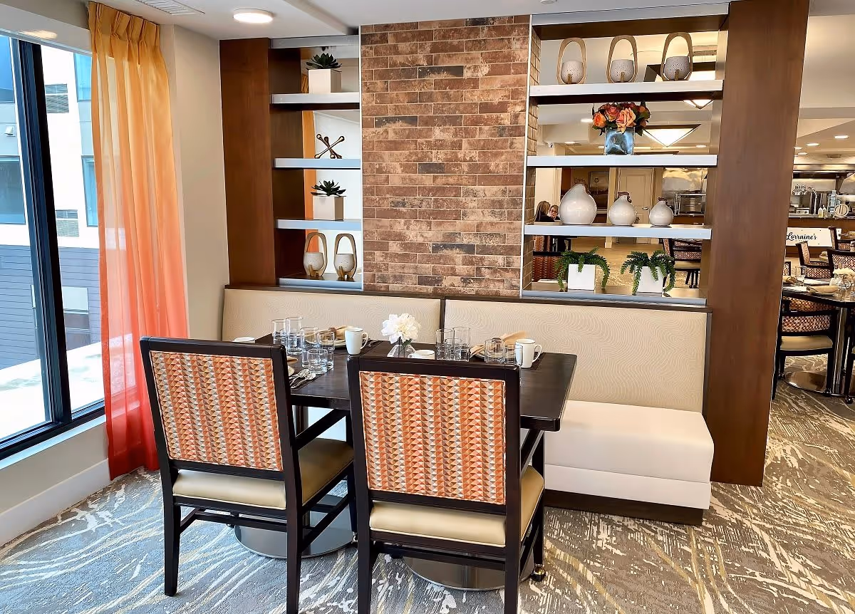A dining nook with a table set for four, upholstered chairs and bench seating, decorative shelving and a large window with orange curtains.