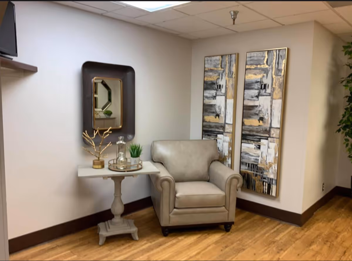 A cozy corner in a room featuring a beige armchair next to a small white side table with decorative items including a gold branch sculpture, a glass jar, and a small potted plant. Above the table is a rectangular mirror with a dark frame. On the adjacent wall, there are two abstract paintings with gold, gray, black, and white tones. The floor is wooden, and there is a green plant partially visible on the right side.