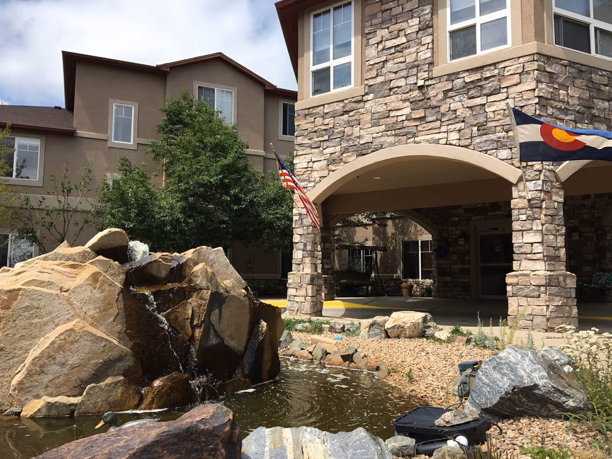 Exterior view of a senior living facility with a stone facade entrance, an American flag and a Colorado state flag displayed, a small pond with rocks and a waterfall feature in the foreground, and trees and windows visible on the building.