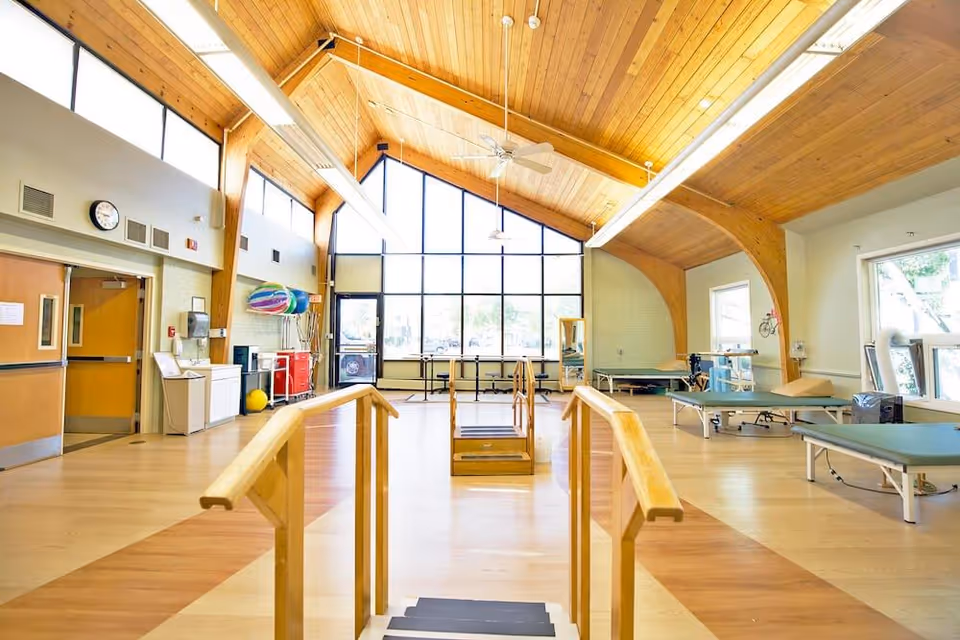 Bright rehabilitation therapy room with parallel walking bars, treatment tables and large vaulted wood ceiling and windows.