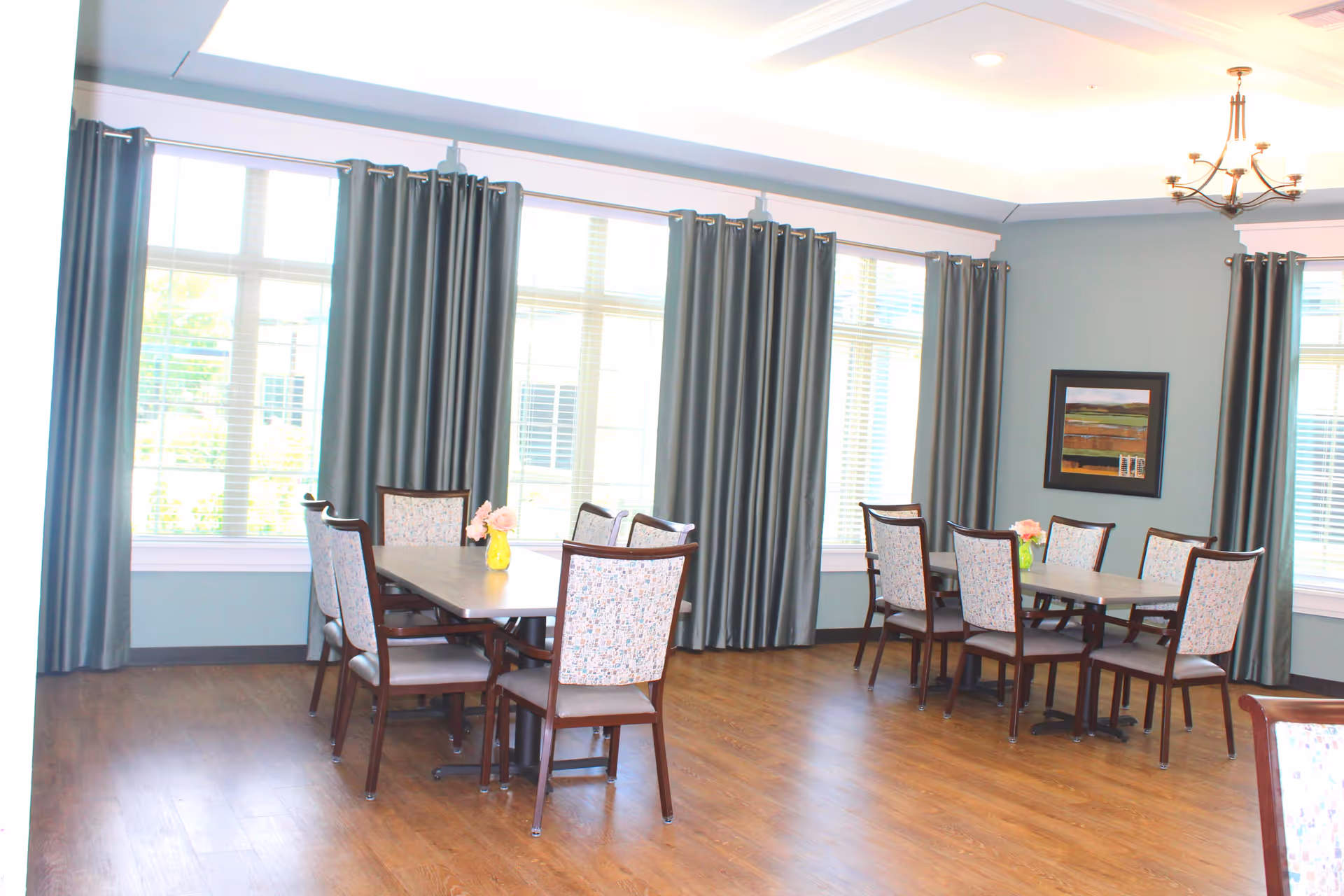 A bright dining room with two rectangular tables, each surrounded by six upholstered chairs. The room has large windows with gray curtains, light blue walls, wooden flooring, a chandelier, and a framed landscape painting on the wall.
