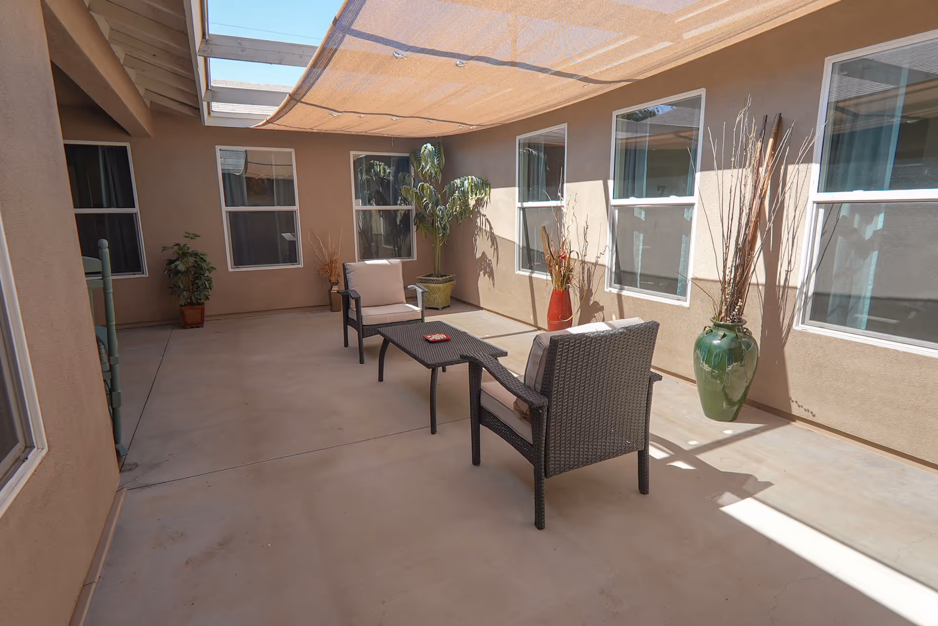 A shaded outdoor patio area with two cushioned wicker chairs and a matching coffee table. The patio is surrounded by beige walls with several windows and decorated with large potted plants and tall vases with dried branches.