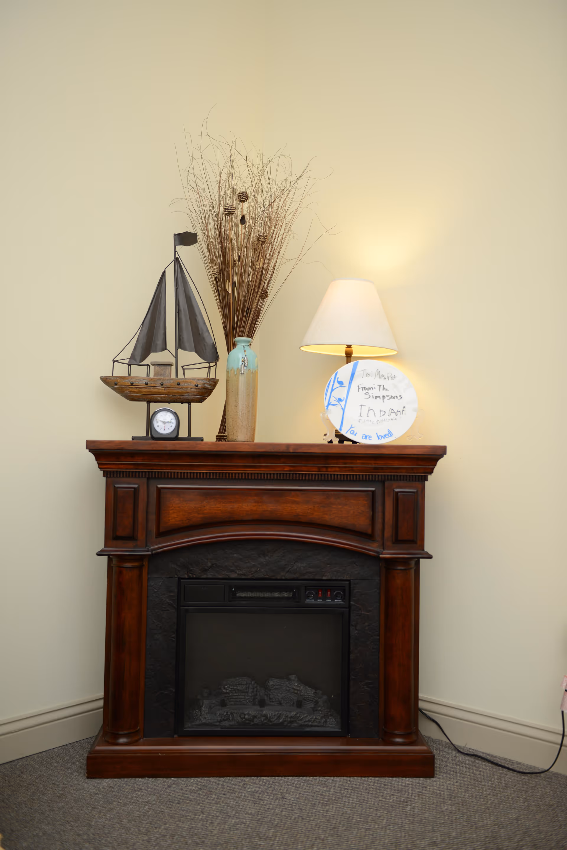 A wooden electric fireplace with a dark finish against a beige wall. On top of the fireplace mantel, there is a decorative model sailboat, a tall vase with dried branches, a table lamp with a white shade, and a round plate with handwritten text. The floor is carpeted in a neutral color.