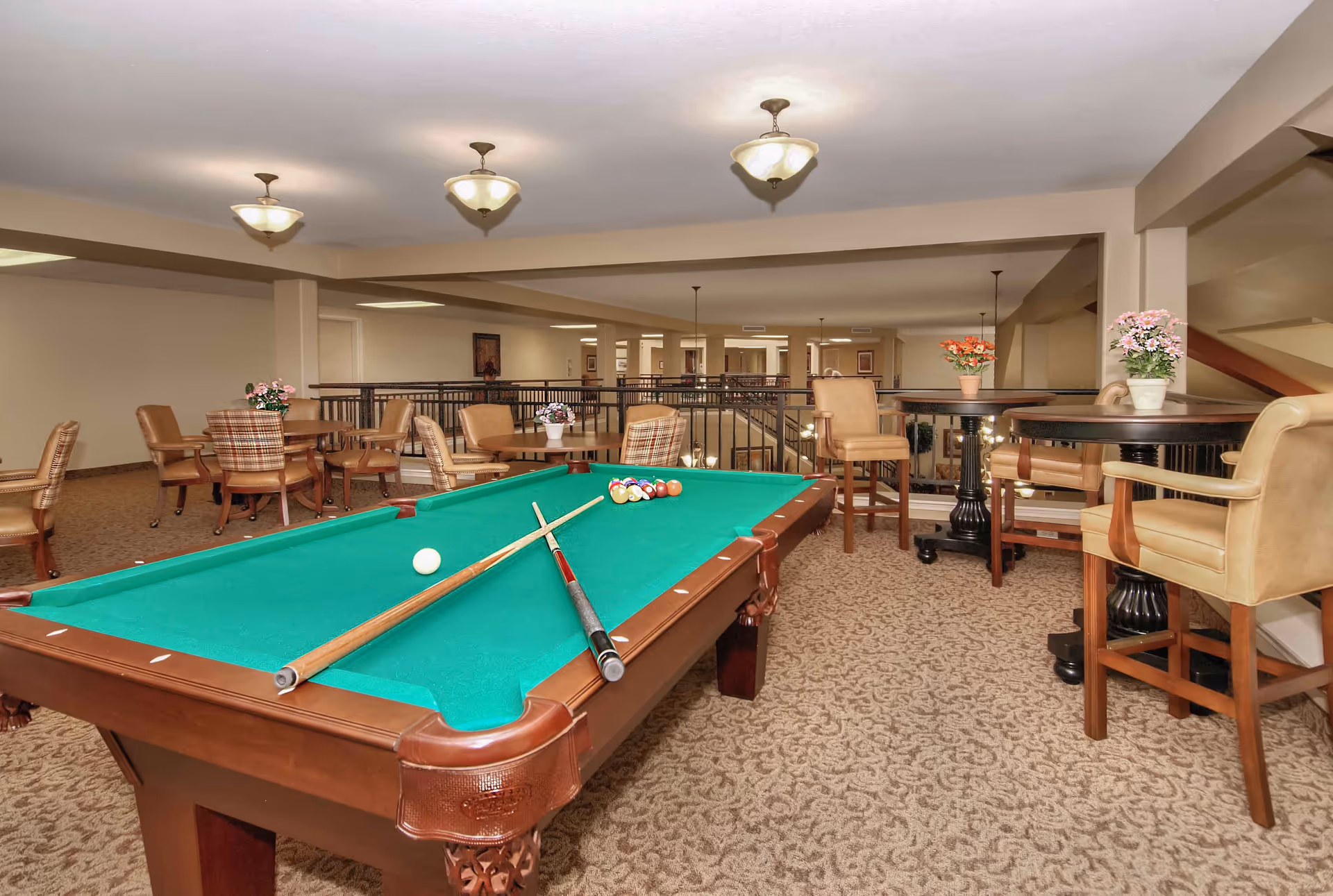 A spacious indoor recreational area featuring a green felt pool table with two cues and a rack of billiard balls. Surrounding the pool table are several seating arrangements including round tables with chairs and high tables with bar stools. The room has beige walls, carpeted floors with a patterned design, and ceiling lights. Potted flowers decorate the tables, and a railing overlooks a lower level in the background.