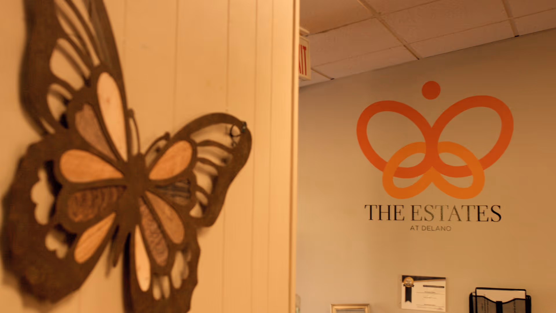 Interior wall with a large decorative butterfly sculpture on the left and a logo on the right that reads 'THE ESTATES AT DELANO' with an orange butterfly-like design above the text.