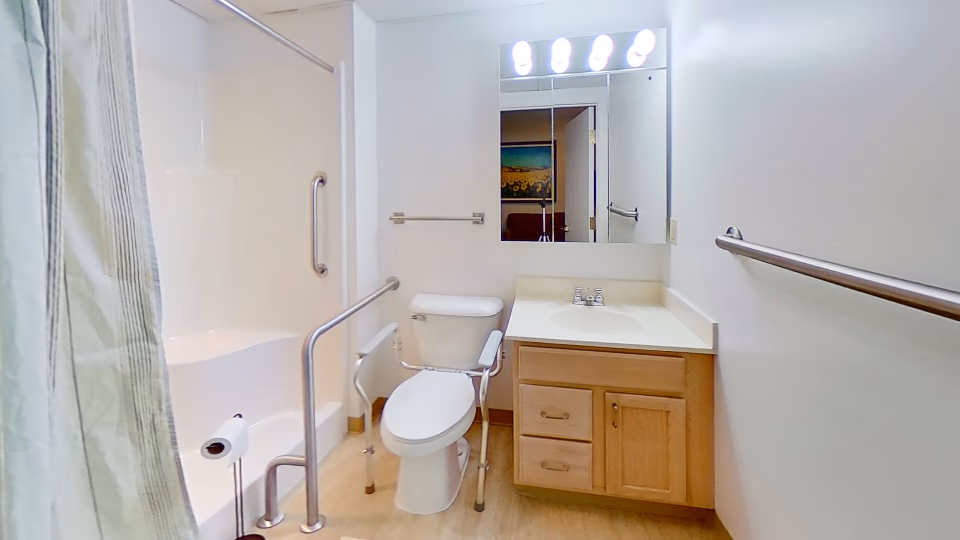 A clean, accessible bathroom with a toilet equipped with safety rails, a bathtub with grab bars, a sink with a wooden cabinet underneath, a large mirror above the sink, and bright overhead lighting. The bathroom has light-colored walls and flooring.