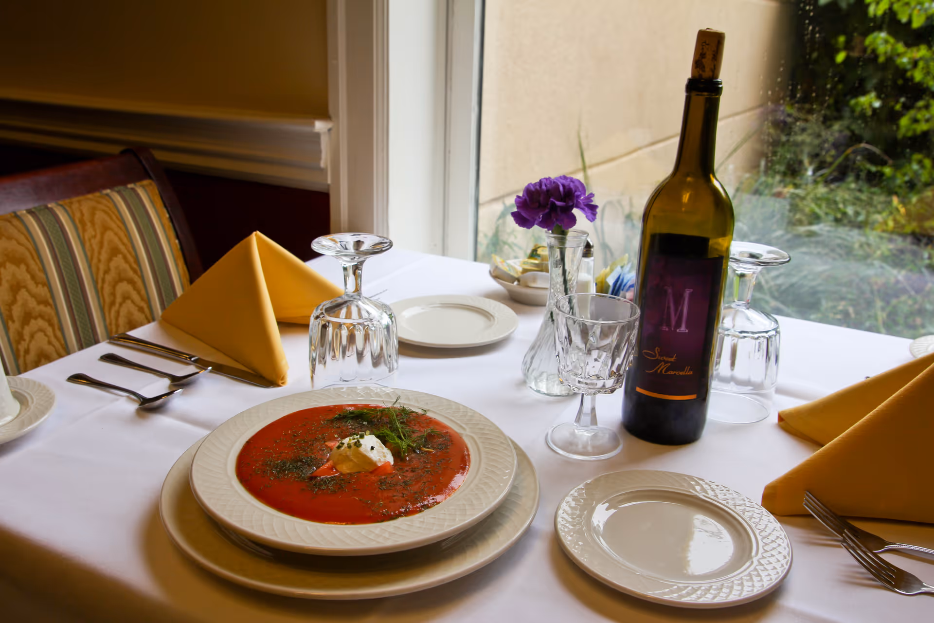 A dining table set with a bowl of tomato soup garnished with cream and herbs, a bottle of wine, a wine glass, two upside-down water glasses, yellow folded napkins, plates, cutlery, and a small vase with a purple flower, next to a window with a view of greenery outside.