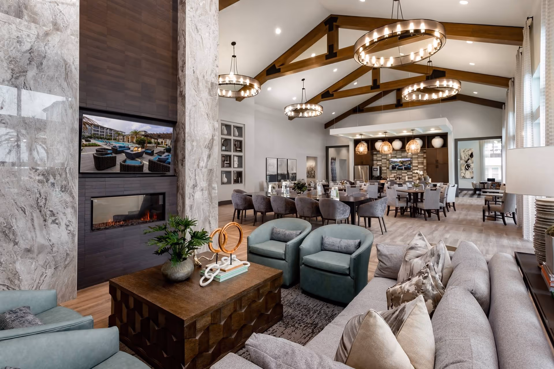 Open communal lounge with sofas and armchairs around a coffee table and fireplace, with dining tables and a kitchen area under exposed wooden beams.