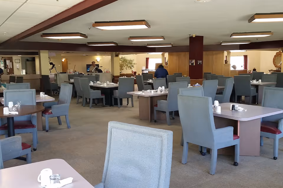 Interior view of a dining room in a senior living facility with multiple tables and chairs arranged neatly. The tables are set with cups, glasses, and napkins. There are a few people in the background near a counter area. The room has beige walls, carpeted floor, and rectangular ceiling lights.