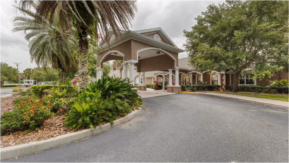 Entrance driveway of a senior living facility with a covered drop-off area supported by white columns. The building is surrounded by lush greenery including palm trees, bushes, and flowering plants under a cloudy sky.