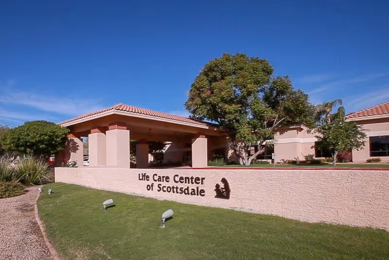 Exterior view of Life Care Center of Scottsdale building with a covered entrance, beige walls, red tile roof, green lawn, and trees under a clear blue sky.