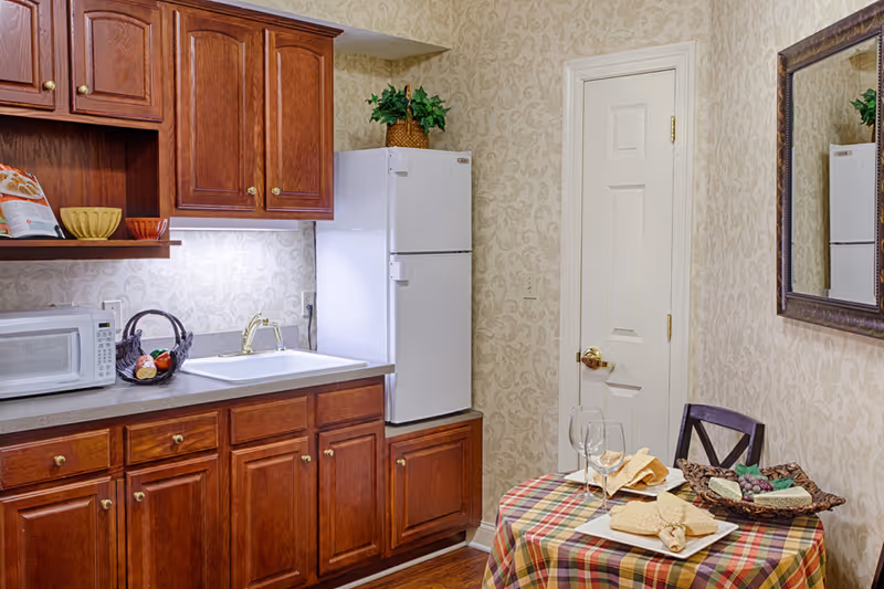 A small kitchen area with wooden cabinets, a white refrigerator, a microwave, and a sink. A round table covered with a plaid tablecloth is set with two wine glasses, napkins, and a basket of snacks. A decorative mirror hangs on the wall above the table, and a potted plant sits on top of the refrigerator.