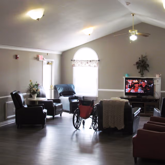 A well-lit living room in an assisted living facility with several armchairs arranged around a television. A wheelchair is positioned near the center of the room. The walls are painted light gray with white trim, and there is a large arched window with a floral valance letting in natural light. A ceiling fan with a light fixture hangs from the ceiling, and a small table with a flower arrangement is visible near the exit door.