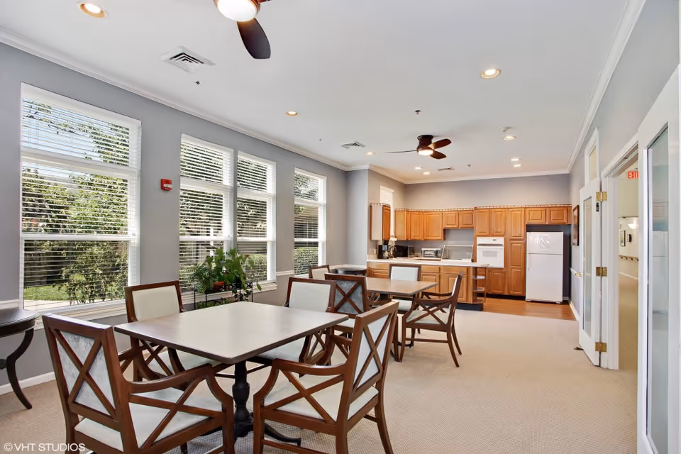 A bright and spacious common area with multiple tables and chairs arranged for dining or socializing. The room features large windows with white blinds allowing natural light to fill the space. In the background, there is a kitchen area with wooden cabinets, a refrigerator, an oven, and a microwave. Ceiling fans and recessed lighting are installed on the white ceiling. The walls are painted light gray, and there is a small plant near the windows.