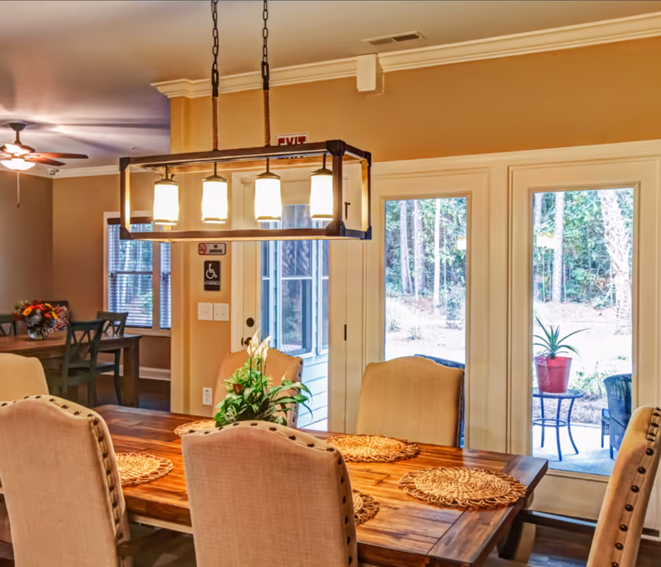 A warm and inviting dining area with a wooden table set with woven placemats and beige upholstered chairs. Above the table hangs a rectangular chandelier with five lights. In the background, there is another dining table with chairs and a ceiling fan with lights. Large glass doors open to an outdoor patio with a small table and potted plant, and a wooded area is visible outside.