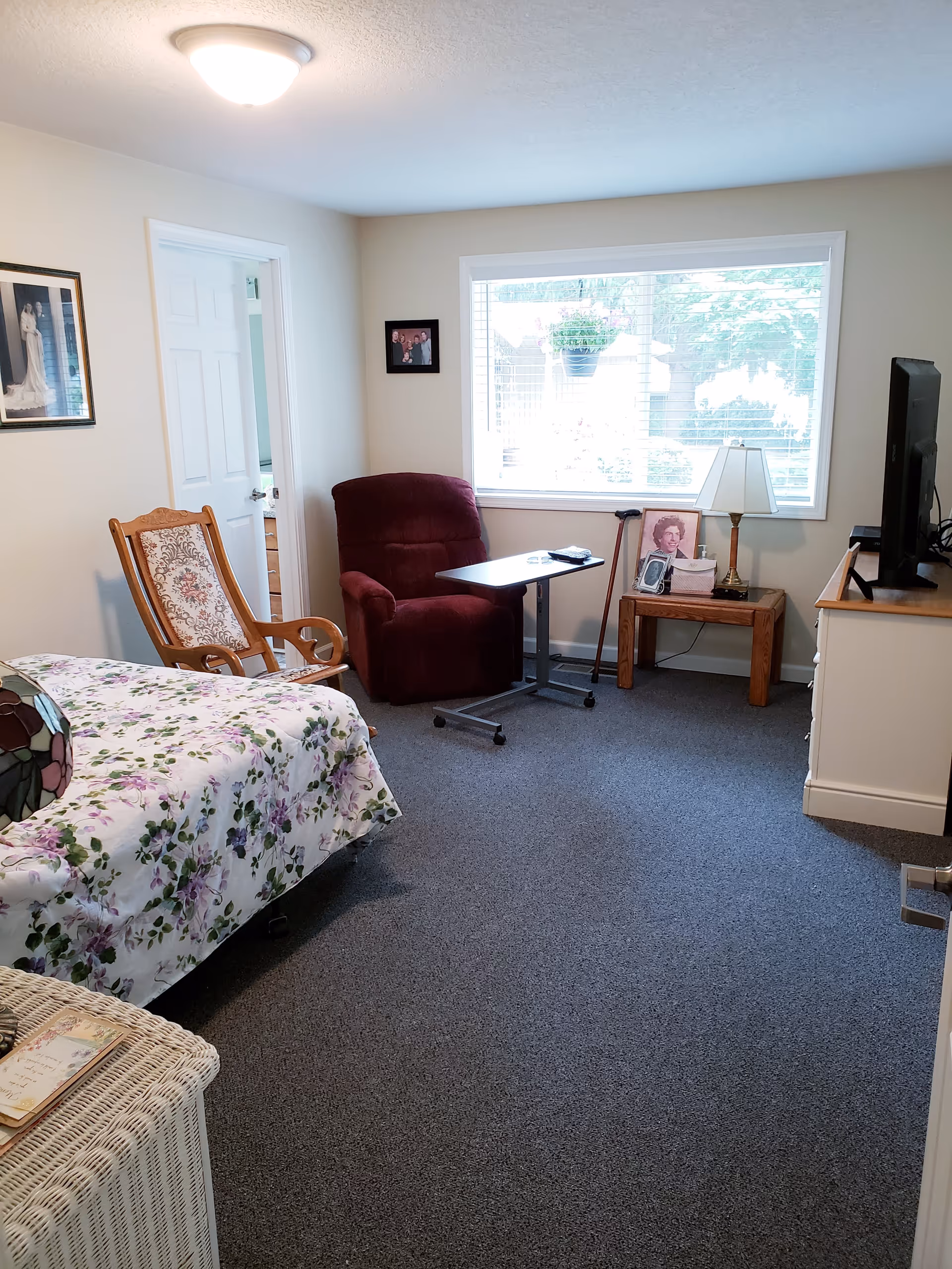 A cozy bedroom in a senior living facility with a floral bedspread on the bed, a wooden rocking chair, a burgundy recliner with an overbed table, a wooden side table with a lamp and framed photos, a large window with blinds showing greenery outside, and a TV on a white dresser.