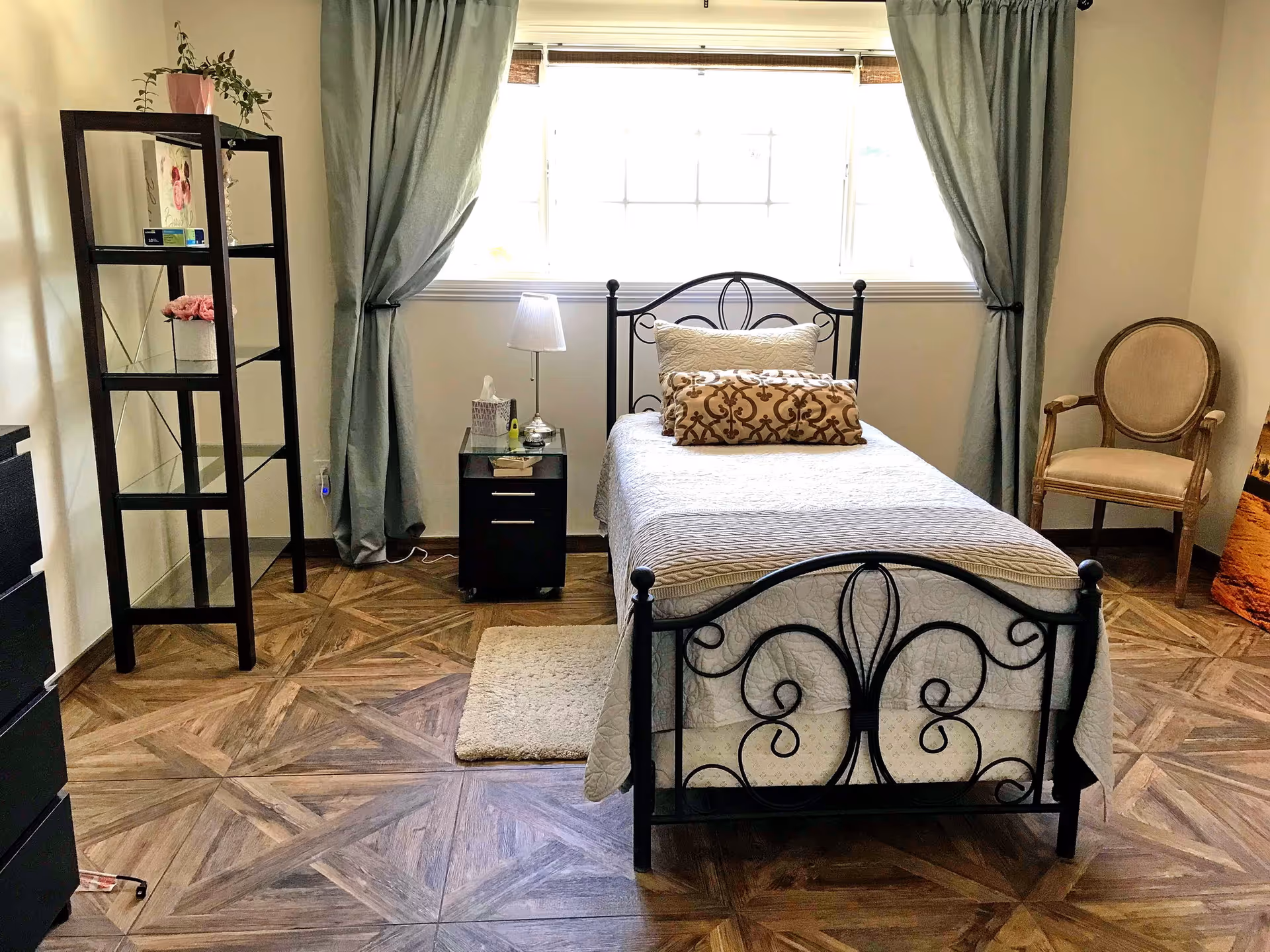 Small bedroom with a single ornate metal bed, nightstand and shelving under a large curtained window.