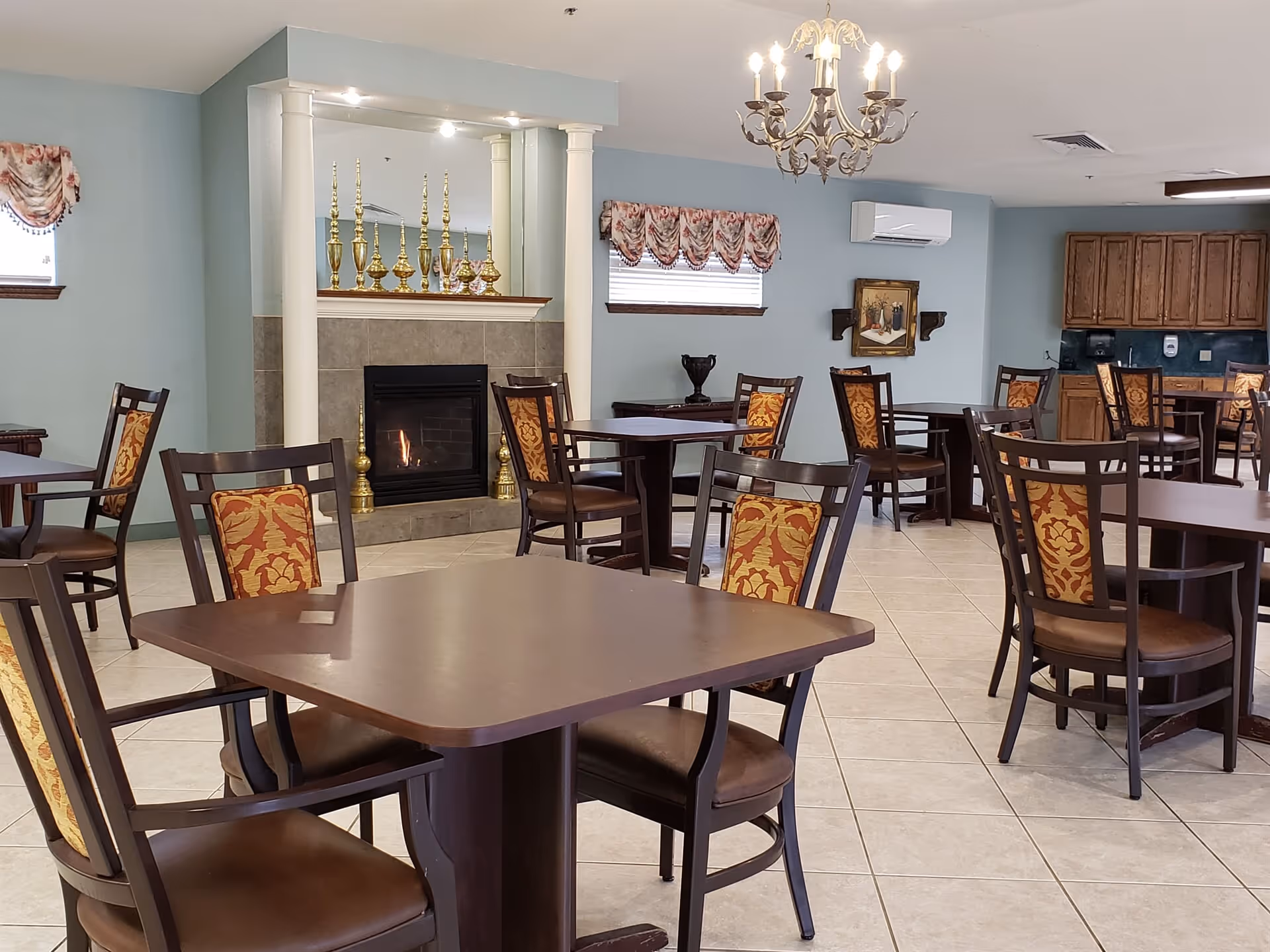 A dining area with multiple dark wood tables and chairs featuring patterned upholstery. The room has light blue walls, a tiled floor, a fireplace with decorative golden candle holders on the mantel, and a chandelier hanging from the ceiling. There are windows with floral valances and wooden cabinets along one wall.