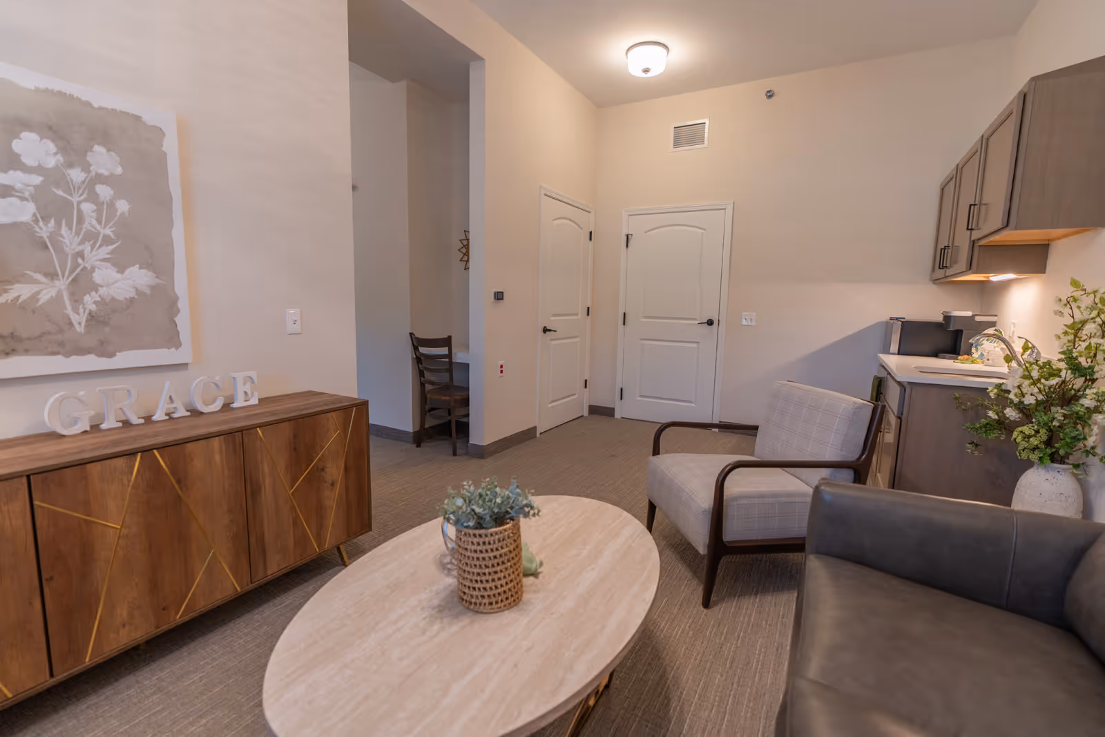 A cozy living room area with chairs, a round coffee table, a wooden sideboard topped with letters spelling "GRACE", a small kitchenette and a doorway.
