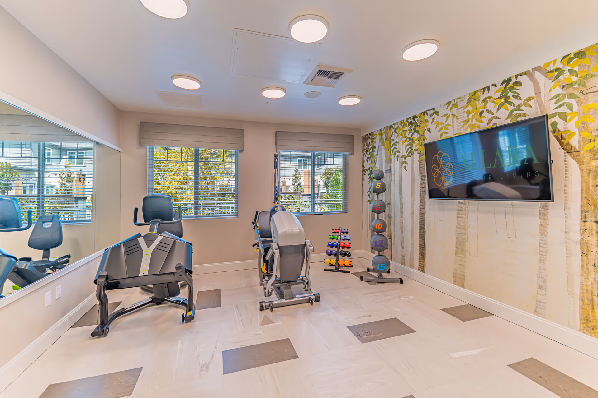 A bright exercise room with two exercise machines, a rack of colorful dumbbells, and a vertical rack of weighted balls. The room has large windows with blinds, a wall-mounted TV displaying the Allara Senior Living logo, and a wall mural featuring trees. The floor has a light-colored tile with darker square accents, and the ceiling has multiple round lights.