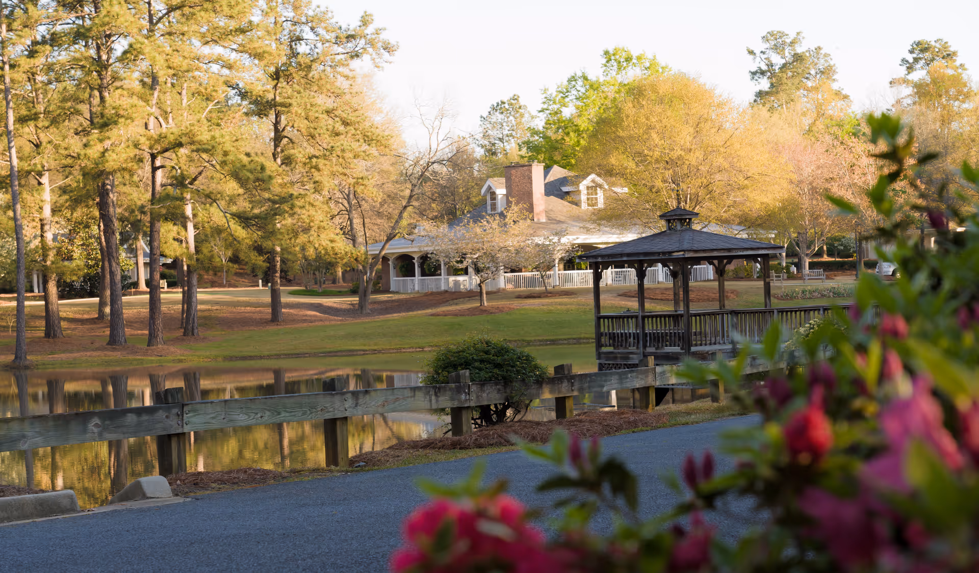 A peaceful outdoor scene at Brandon Wilde featuring a wooden gazebo on a small pier extending over a pond, surrounded by trees with autumn foliage and a large house with a wraparound porch in the background. Pink flowers are visible in the foreground.
