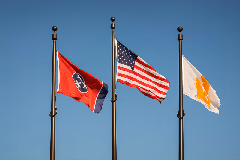 Three flags flying on flagpoles against a clear blue sky. From left to right: the Tennessee state flag, the United States flag, and a white flag with an orange emblem.