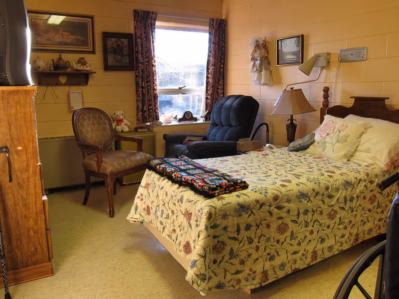 A cozy bedroom with a single bed covered in floral bedding, a recliner and armchair by a window and a bedside lamp.
