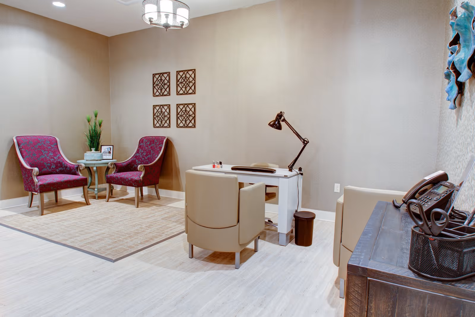 A cozy interior room with two purple upholstered armchairs and a small round table with a plant and a framed sign. Across from them is a white desk with a beige chair, a black desk lamp, and nail polish bottles on top. A wooden cabinet with a telephone and office supplies is visible on the right side. The walls are beige with decorative wall art, and the floor is light-colored wood with a beige area rug.