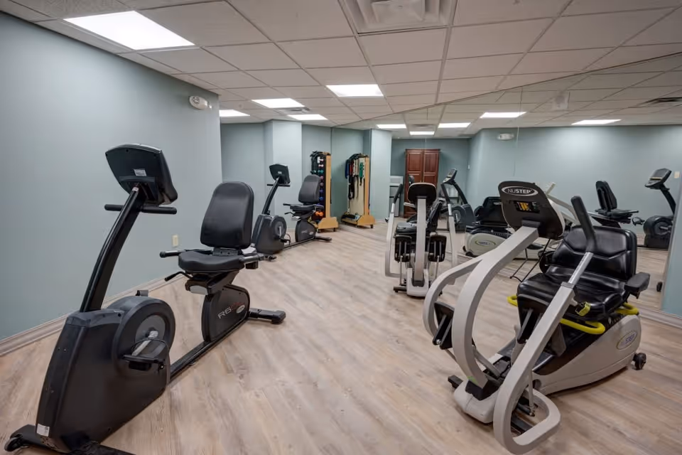 A fitness room with several exercise machines including recumbent bikes and other cardio equipment. The room has light wood flooring, light blue walls, a drop ceiling with fluorescent lighting, and mirrors on one wall. There is also a rack with various weights and exercise bands in the background.