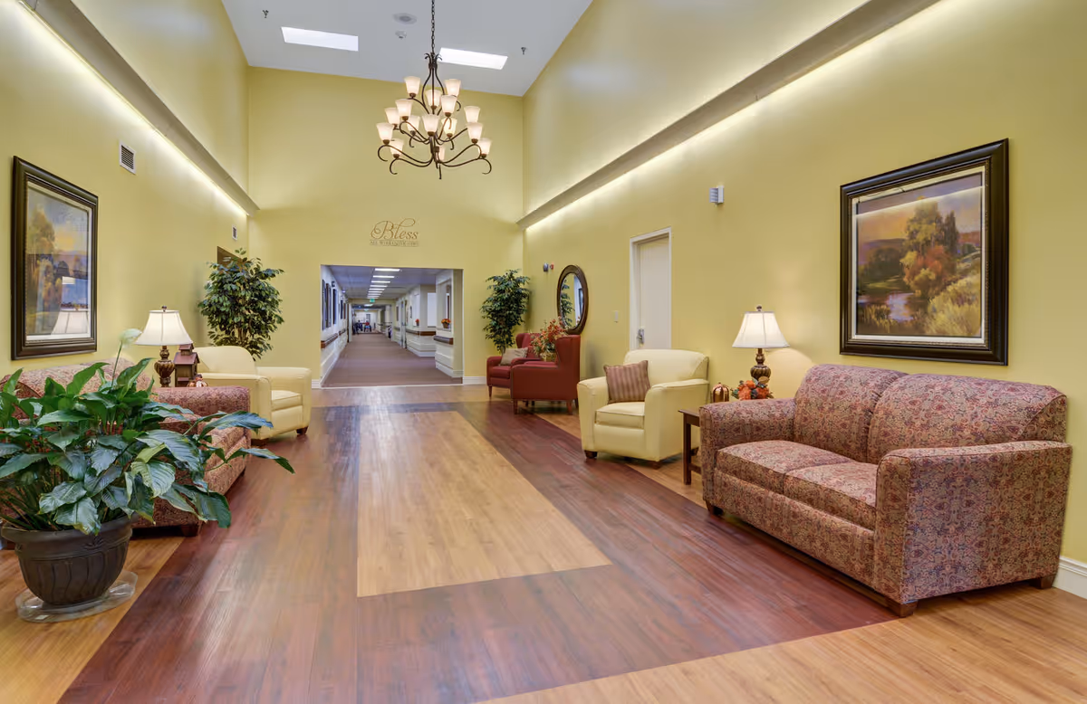 A bright communal lounge with sofas, armchairs, potted plants, framed art, and a long hallway leading deeper into the facility.
