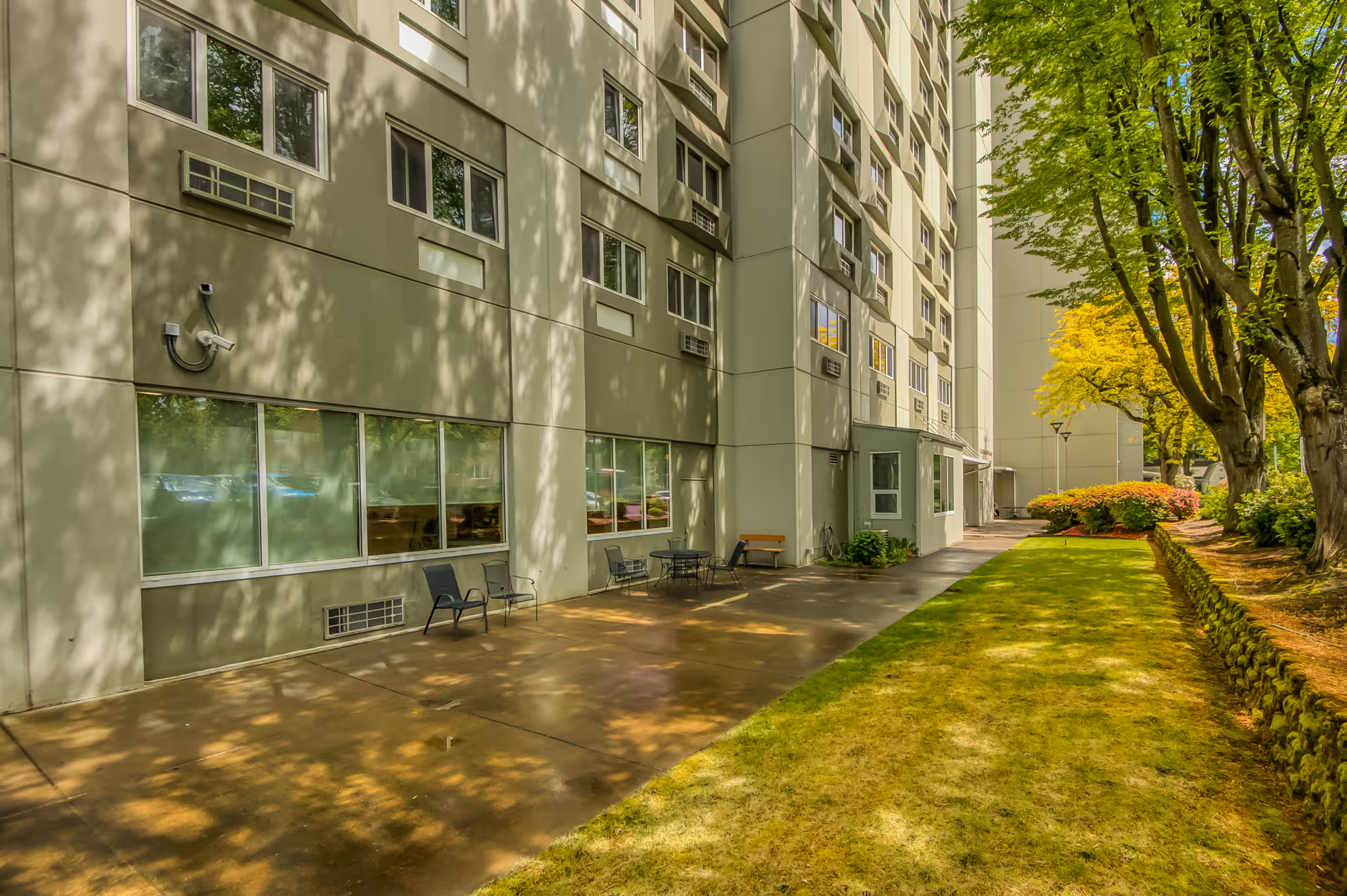 Outdoor view of a senior living facility with a concrete patio area featuring chairs and a table, adjacent to a tall building with multiple windows. The scene includes a grassy lawn bordered by trees and shrubs, with sunlight casting shadows on the building and ground.