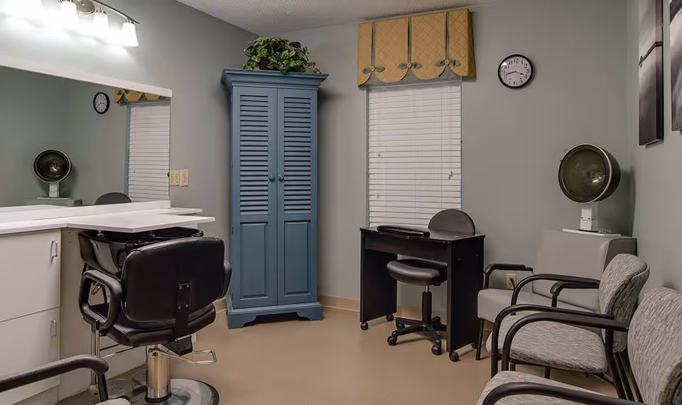 A small salon or beauty room with a black salon chair in front of a white counter and mirror, a blue cabinet with a plant on top, a black desk with a rolling chair, and three waiting chairs along the wall. There is a hair dryer hood on a stand near the waiting chairs, a window with closed blinds and a yellow valance, and a clock on the wall.