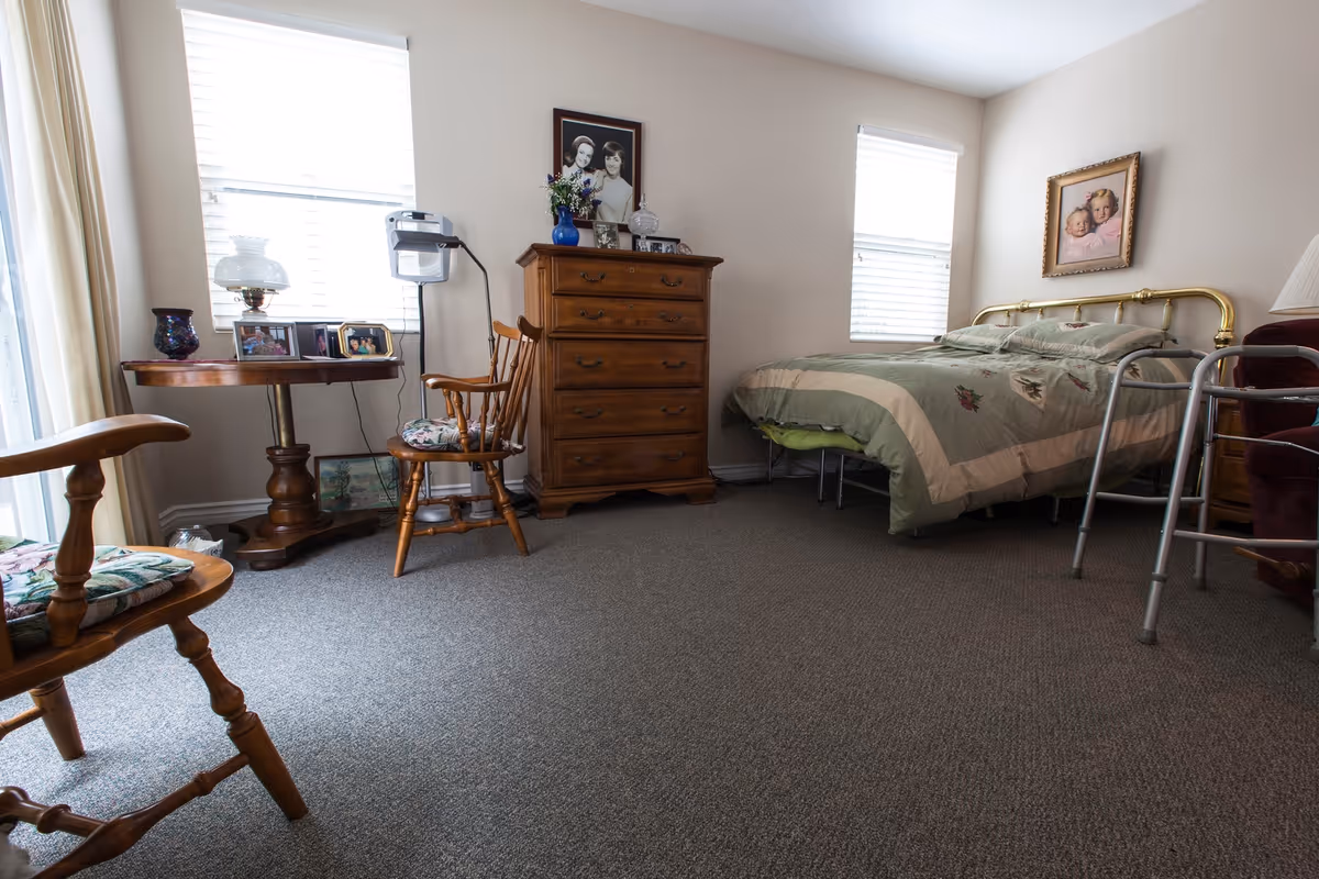Sunny bedroom with a brass bed, wooden dresser and chairs, a side table with photos, and a walker near the bed.