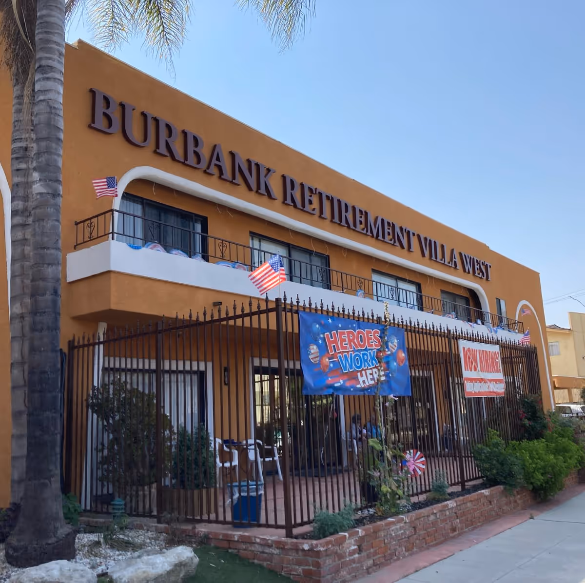 Exterior view of a two-story building with a sign reading 'BURBANK RETIREMENT VILLA WEST'. The building is orange with white trim and has a fenced patio area decorated with American flags and banners, one of which says 'HEROES WORK HERE'. There are some plants and a palm tree visible in front of the building under a clear blue sky.