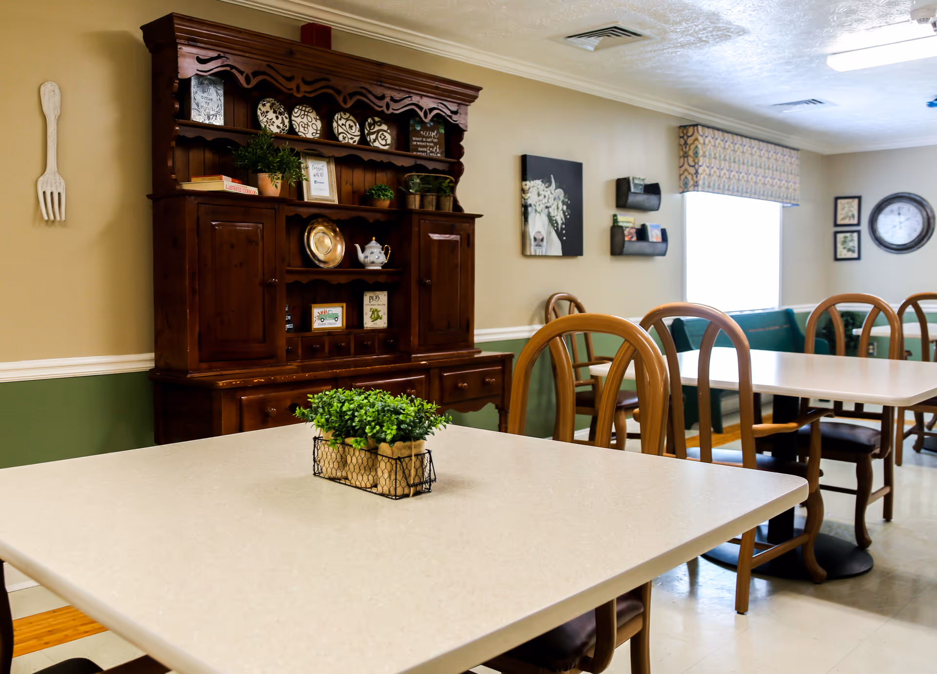 A bright dining room with tables and wooden chairs, a decorative wooden hutch against the wall, and a small potted centerpiece on the nearest table.