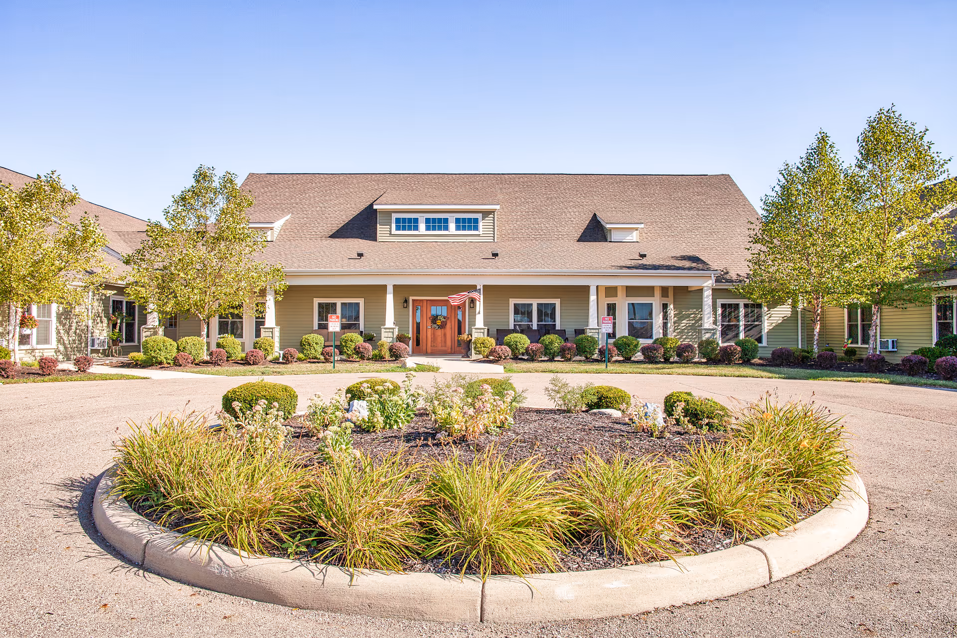Front exterior view of a single-story senior living facility building with a circular driveway and landscaped garden in the center. The building has a beige exterior with a brown roof, several windows, and a central entrance with double wooden doors and an American flag above.