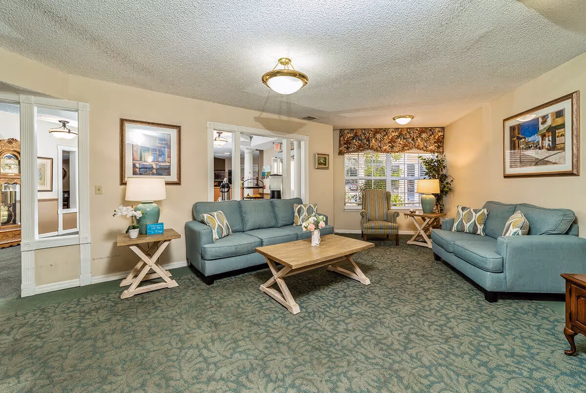 A cozy living room with two teal sofas, a wooden coffee table, and two side tables with lamps. There is a striped armchair near a window with floral valance, framed artwork on the walls, and a green patterned carpet. The room has a warm and inviting atmosphere.