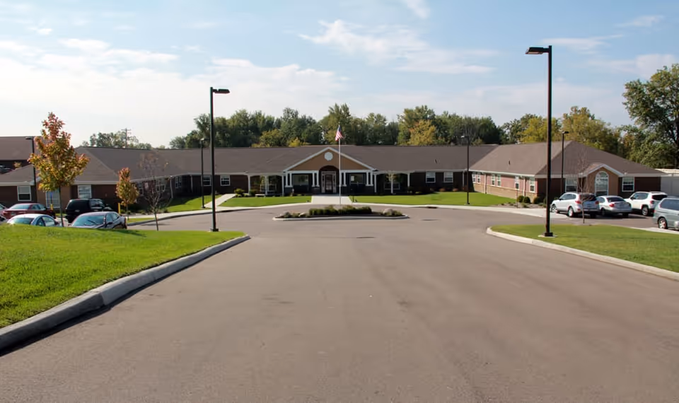 Front exterior view of a single-story senior living facility building with a central entrance, surrounded by a paved driveway, parking spaces with cars, green lawns, and trees under a partly cloudy sky.