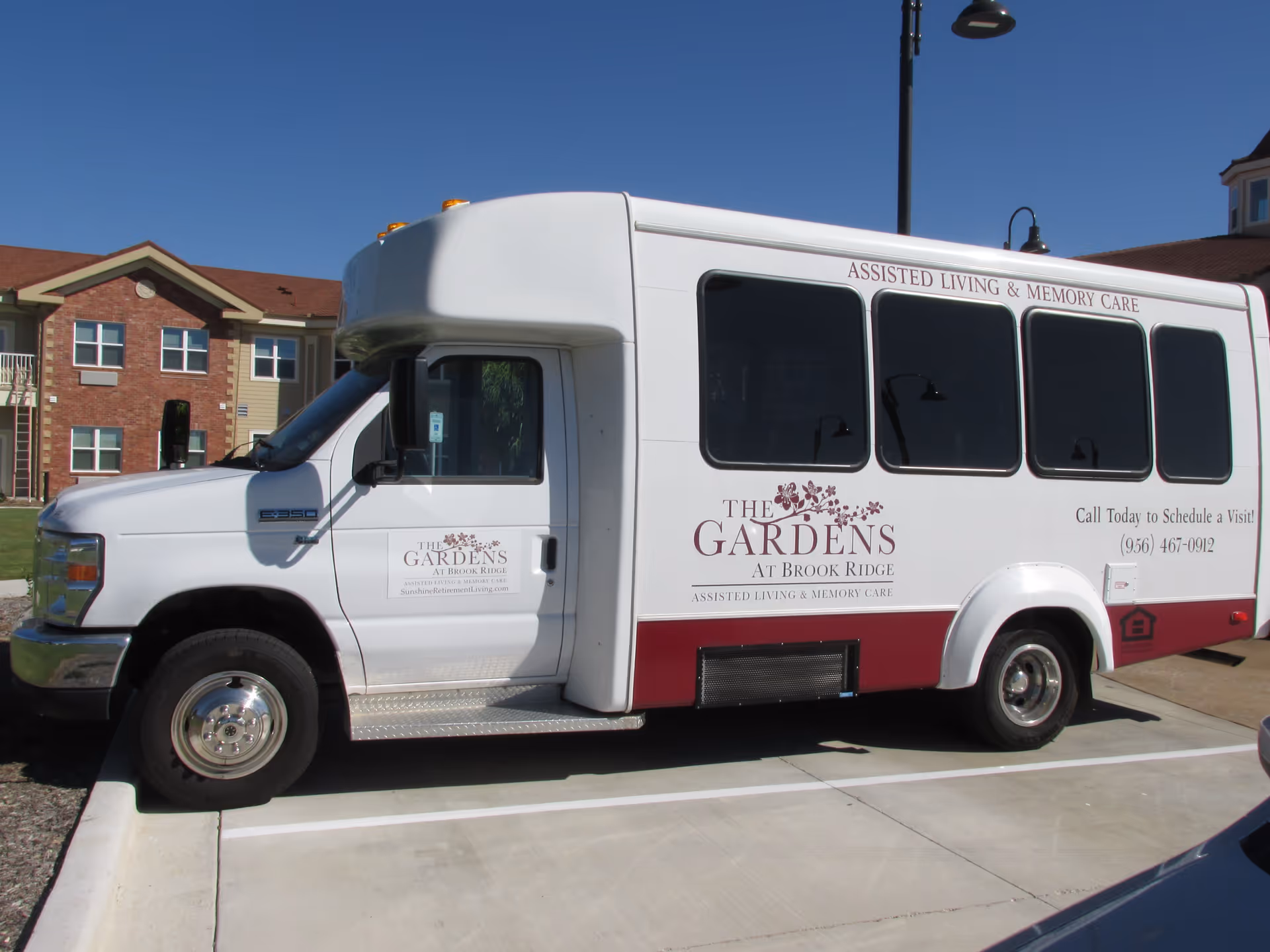 A white shuttle bus parked in a parking lot in front of a residential building. The bus has signage for The Gardens at Brook Ridge Assisted Living & Memory Care, including contact information to schedule a visit. The building in the background has red brick and beige siding with multiple windows.