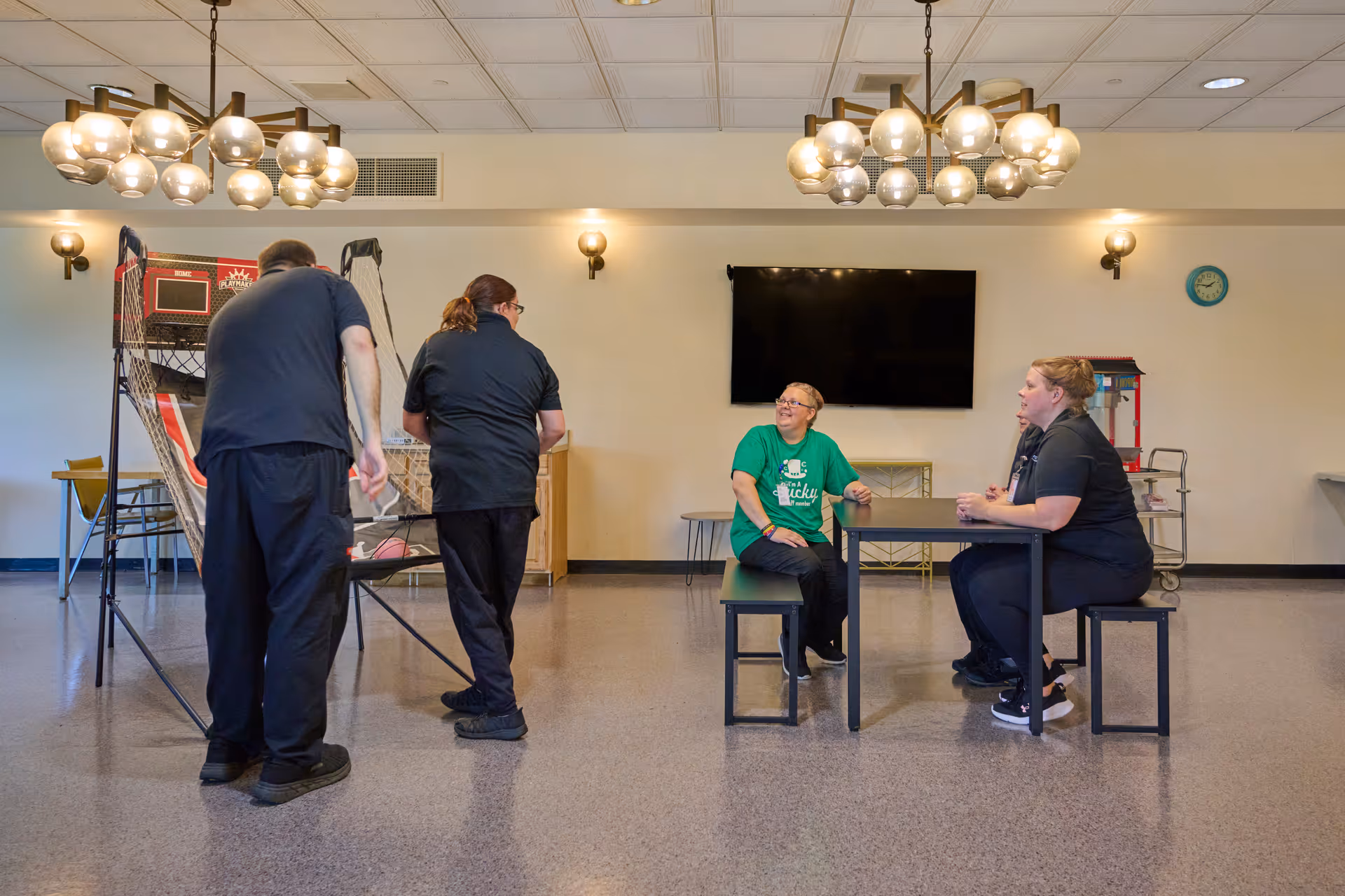 A common room in a senior living facility with four people. Two people are playing a basketball arcade game on the left side, while two others sit at a table on the right side. The room has modern light fixtures, a wall-mounted TV, a clock, and a popcorn machine in the background.