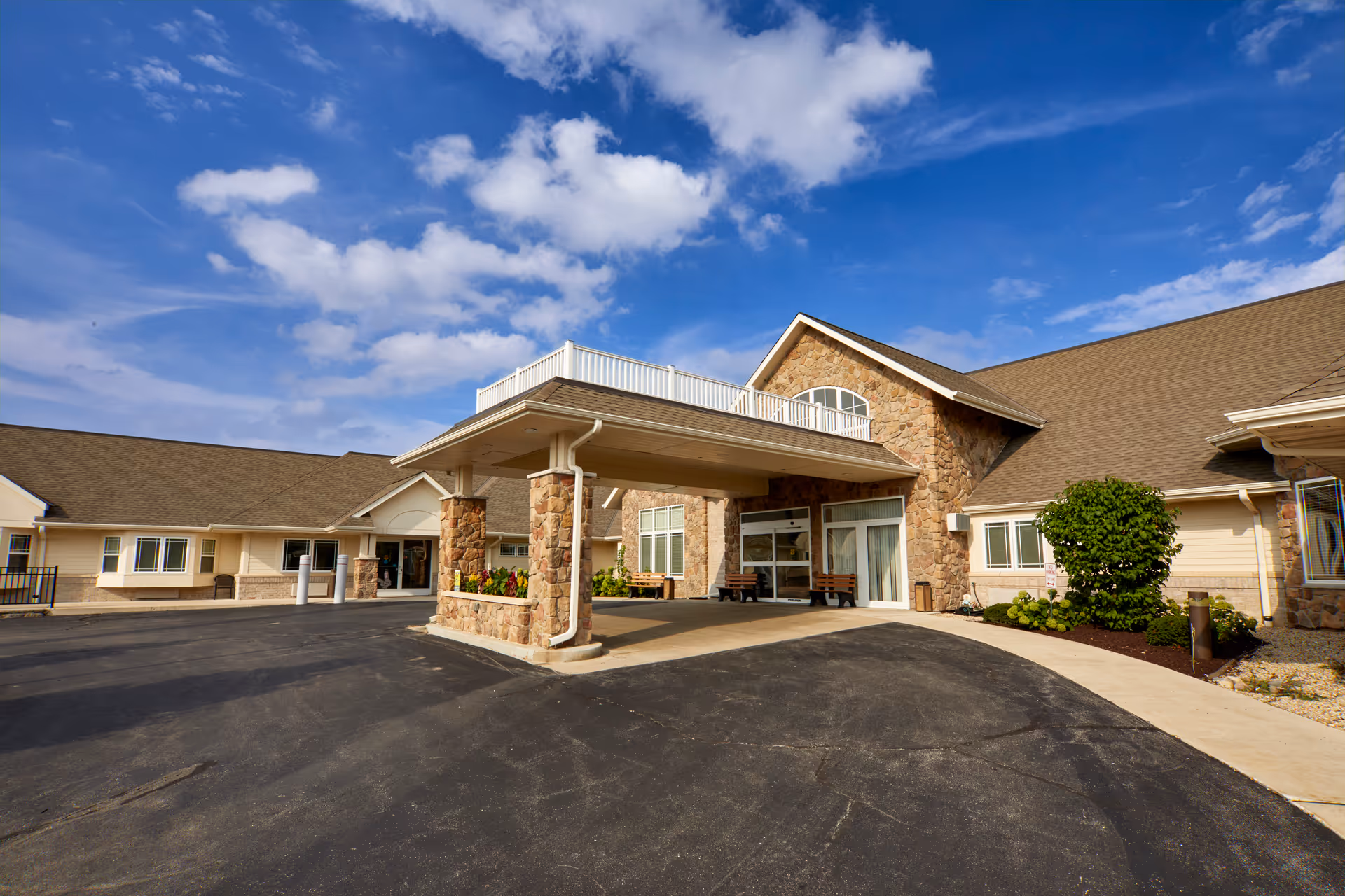 Exterior view of Autumn Lake Healthcare at Greenfield showing the main entrance with a covered drop-off area supported by stone pillars. The building has a stone and beige siding facade with a sloped roof under a partly cloudy blue sky.