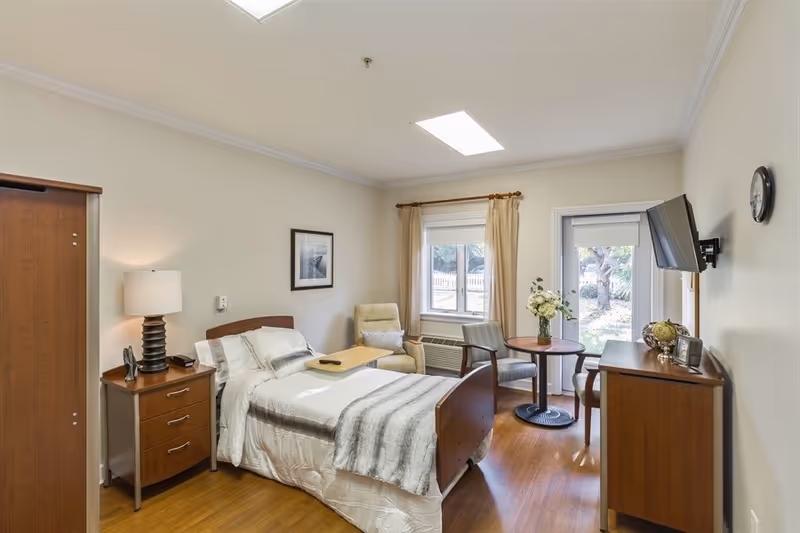 A well-lit bedroom in a senior living facility featuring a single bed with white and gray bedding, a wooden nightstand with a lamp, a comfortable armchair, a small round table with two chairs, a wall-mounted TV, and a window and door leading outside with beige curtains.